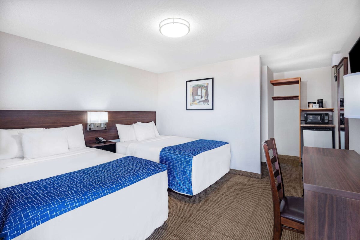 Une chambre d’hôtel avec deux lits, un bureau, une chaise, un four à micro-ondes et une étagère avec divers objets.
