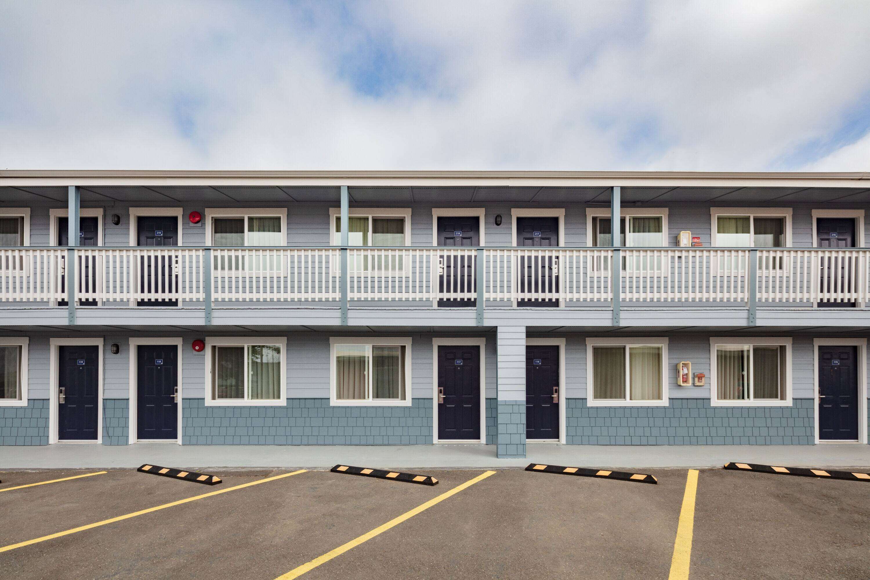 A multi-story motel with a balcony on the second floor, individual rooms with doors, and a parking area with marked spaces.