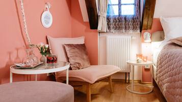 A cozy bedroom with a pink theme, featuring a bed, a chair, a small table with a vase and candles, and a window with lace curtains.