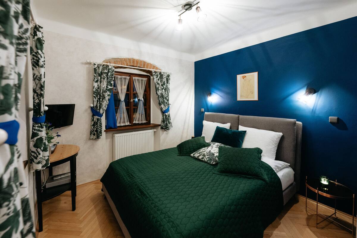 Ein Schlafzimmer mit einem Bett, einem Nachttisch, einem Fenster mit Vorhängen, einer Wandleuchte und einem gerahmten Bild.