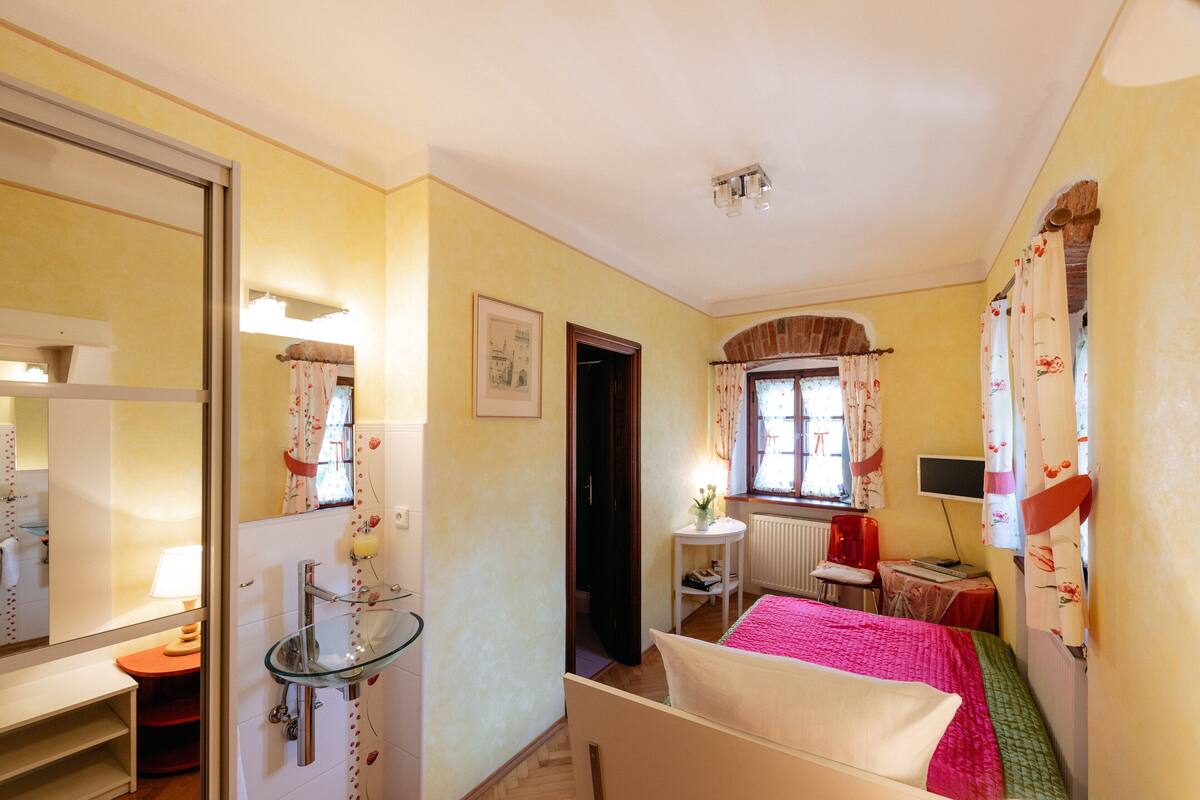 Ein Schlafzimmer mit einem Bett, einem Nachttisch, einem Spiegel, einem Fenster mit Vorhängen, einer Heizung und einem kleinen Tisch mit einem Stuhl.