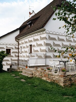 Un edificio blanco con fachada decorativa, una escalera de piedra y un pequeño jardín con sillas blancas y plantas en macetas.