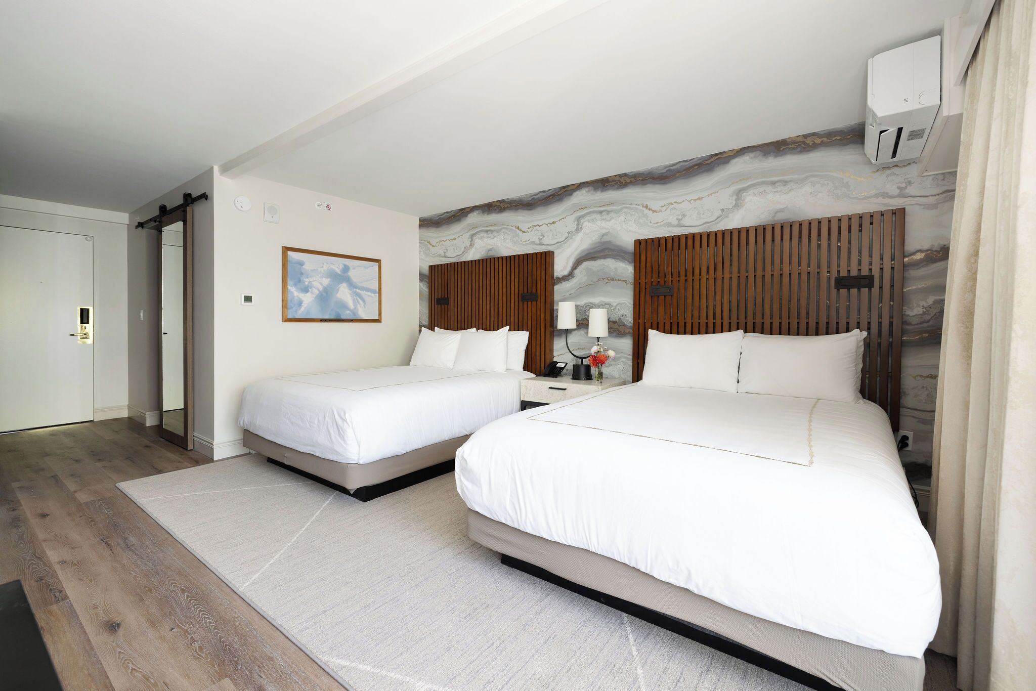 A hotel room with two beds, a marble wall, wooden headboard panels, a mirror, a bedside table with a lamp, and an air conditioning unit.