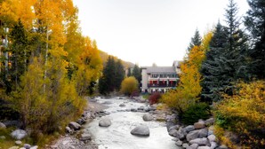 A river with rocks and trees in autumn colors, a building with a red awning, and a mountain in the background. - Sitzmark Vail (Vail)