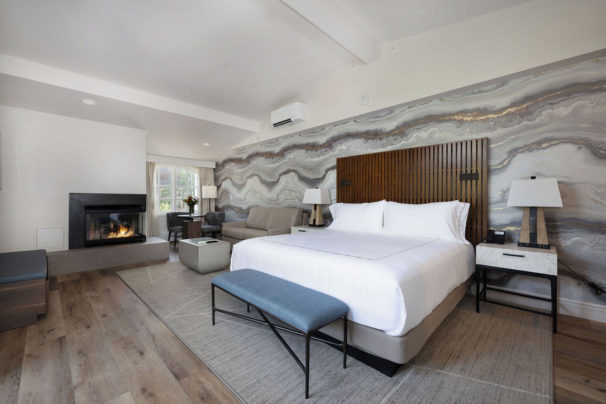 A modern bedroom with a large bed, a fireplace, a bench, and a marble wall.