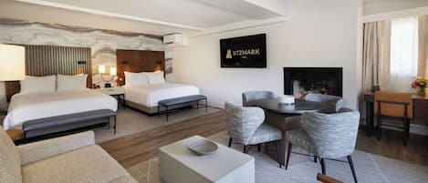 A modern hotel room with two beds, a dining area, and a fireplace.