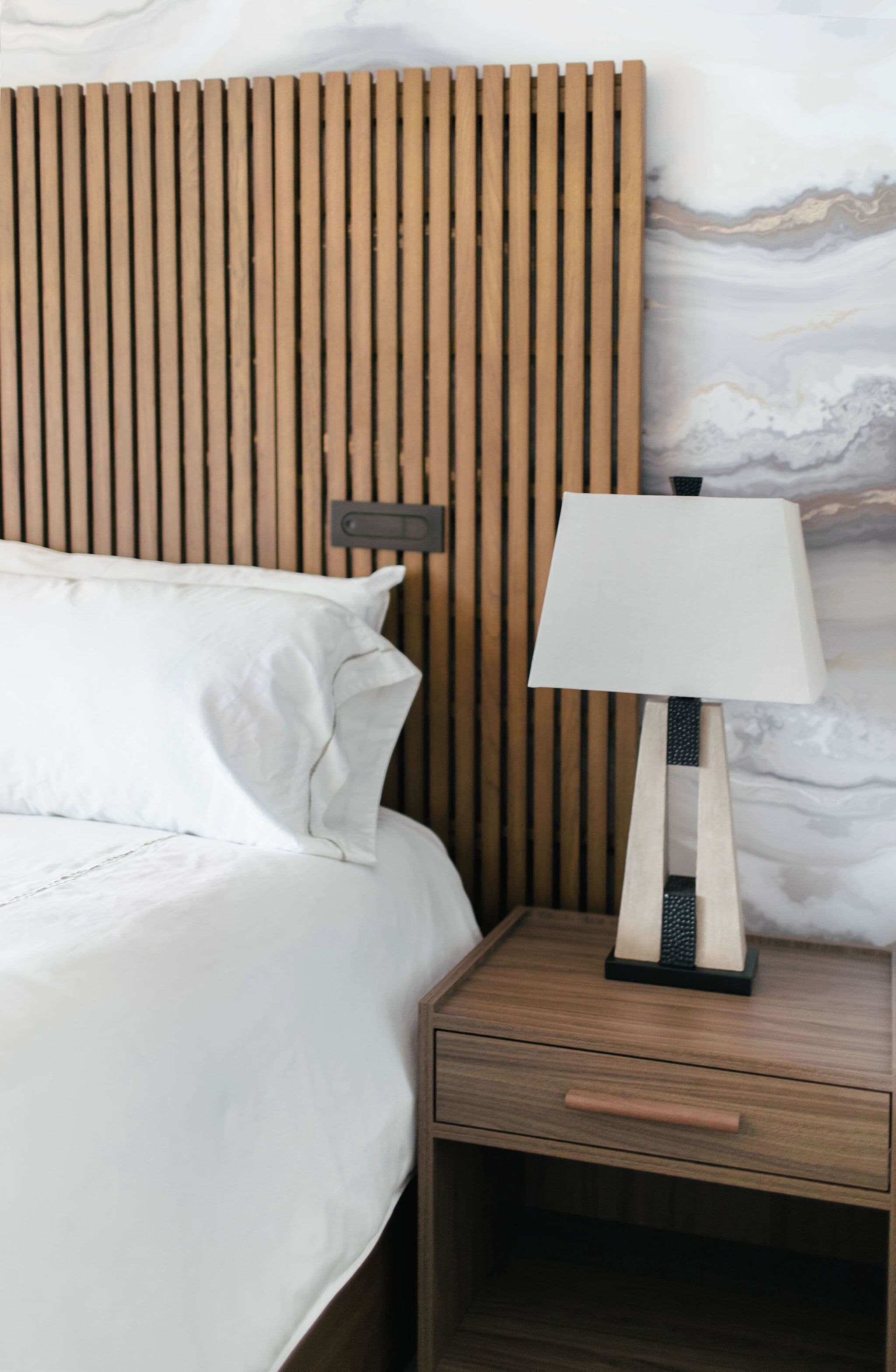 A bed with white bedding, a wooden headboard, a bedside lamp, and a nightstand with a drawer.