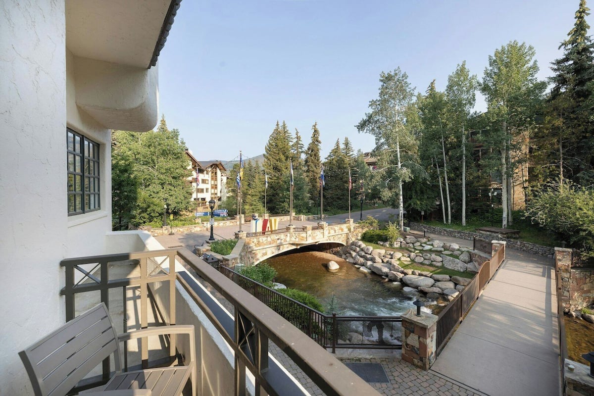 A balcony with a stone bridge over a river, surrounded by trees and buildings.