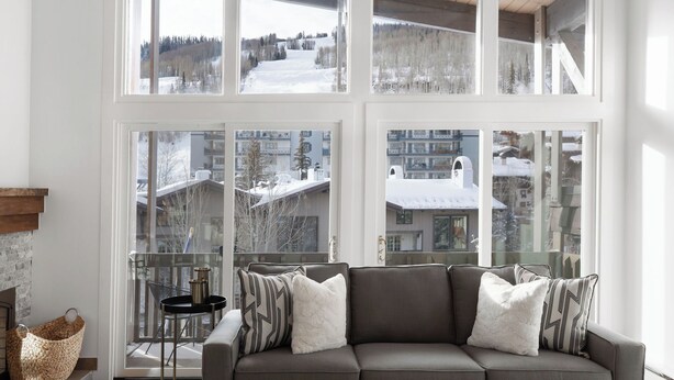 A modern living room with a grey sofa, patterned cushions, and a glass table. Large windows offer a view of a snowy landscape and buildings. - Sitzmark Vail (Vail)