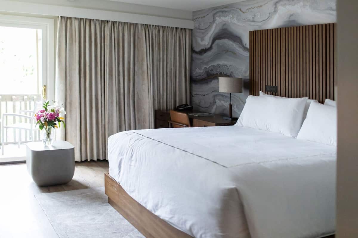 A modern hotel room with a large bed, a desk, a chair, a lamp, and a vase with flowers.