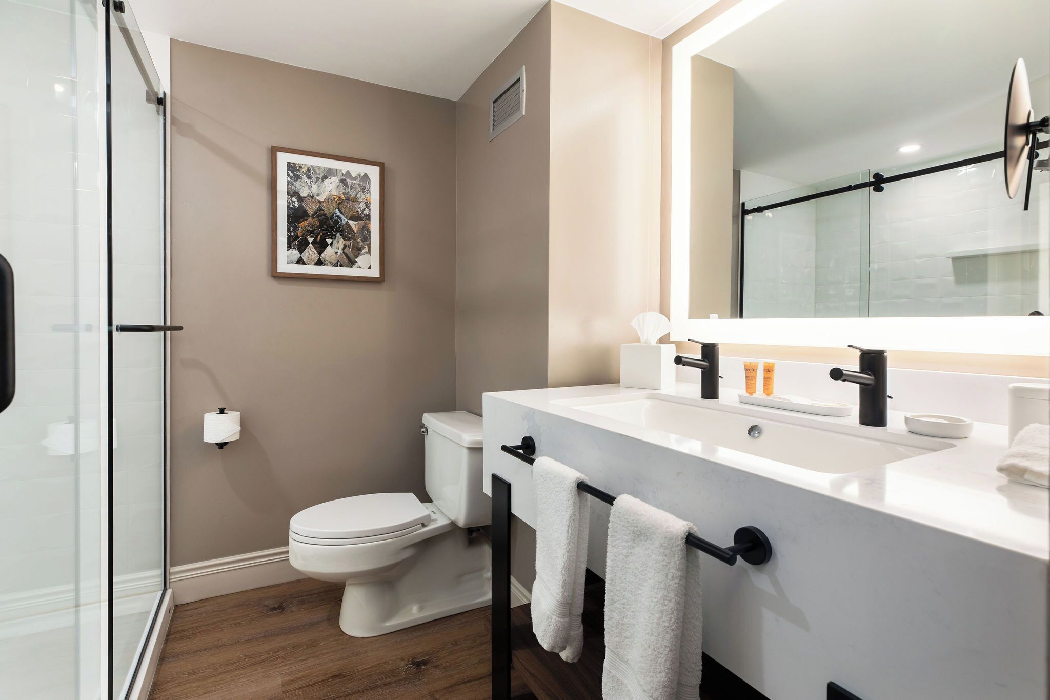 Un baño moderno con cabina de ducha de vidrio, inodoro, lavabo con espejo y un cuadro en la pared.