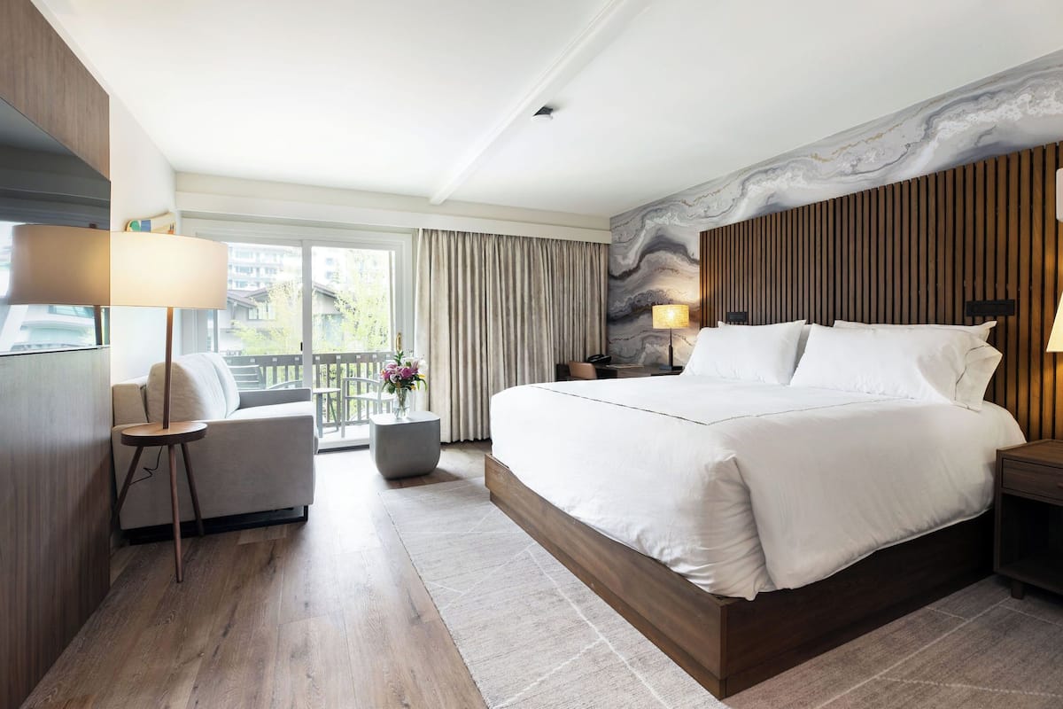 A modern hotel room with a large bed, a sofa, a small table with a lamp, and a view of the outdoors.