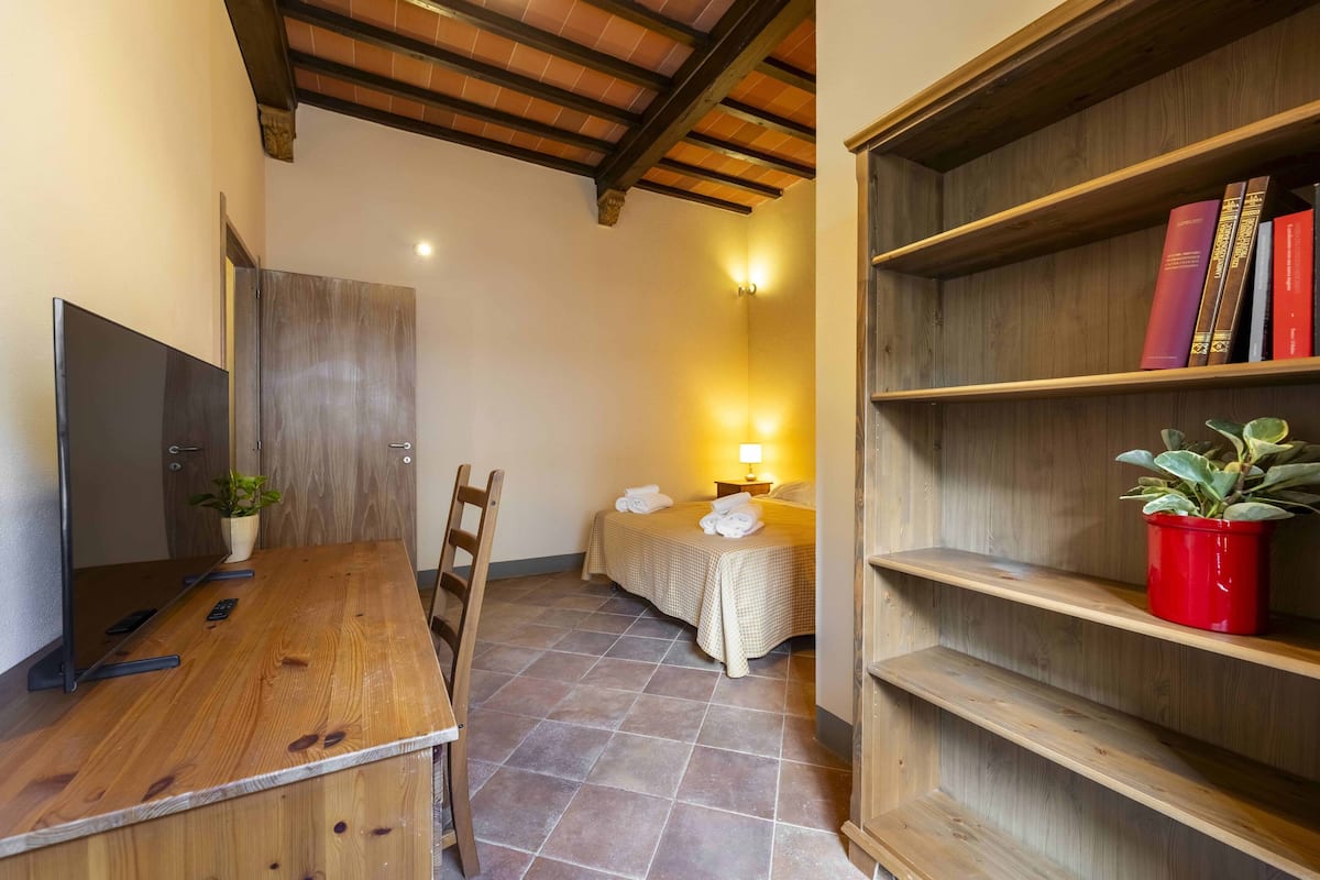 A cozy room with a wooden desk, a bed with a checkered bedspread, a bookshelf with books, and a potted plant.