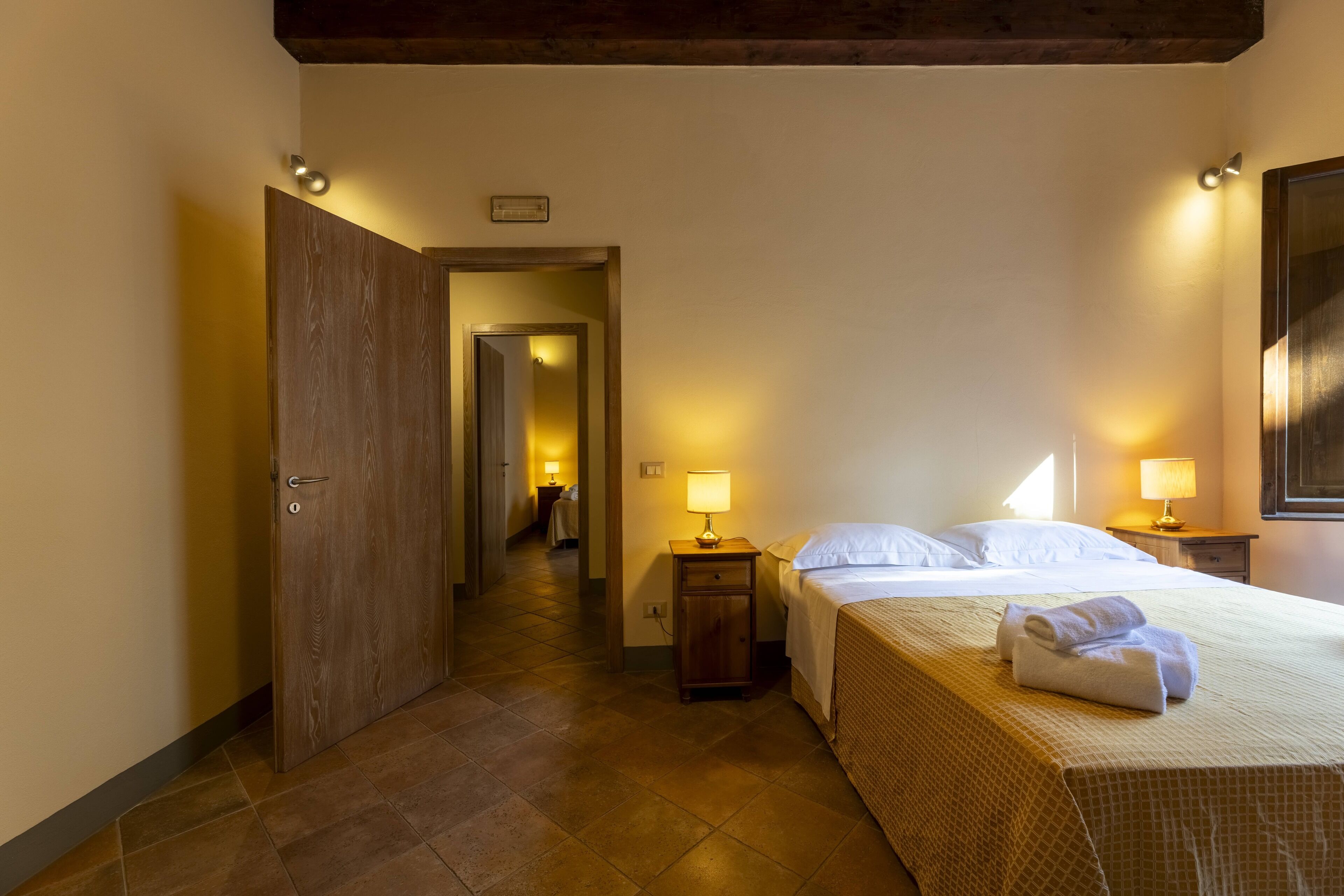 Una habitación de hotel con una cama, lámparas de noche, una puerta de madera y un pasillo que conduce a otra habitación.