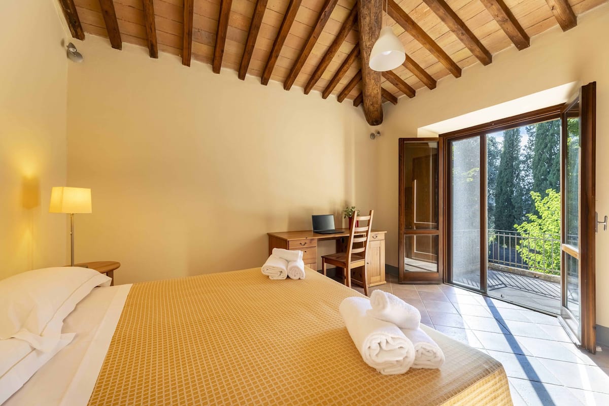 A bedroom with a bed, a desk, a chair, and a balcony with a view of trees.