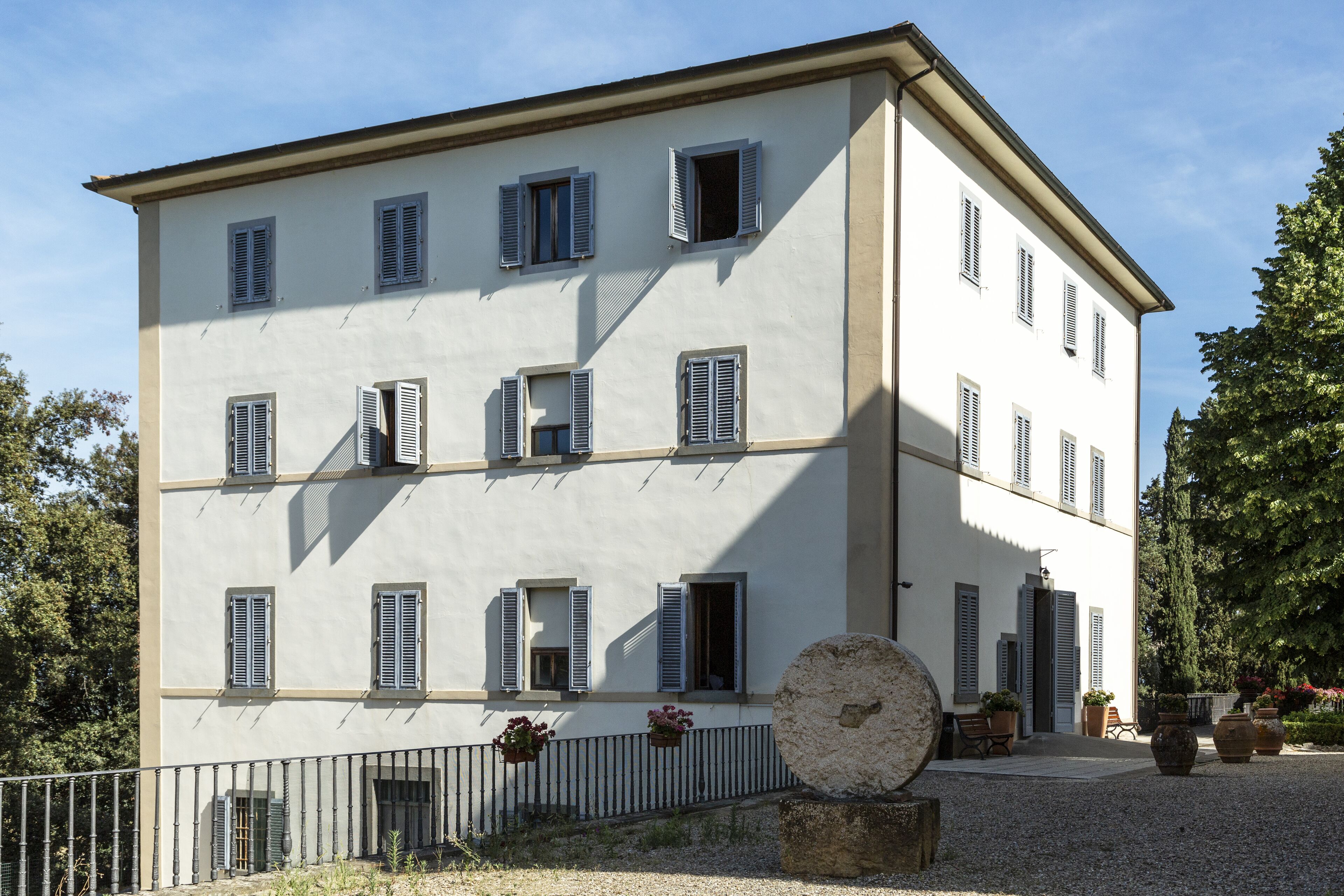 Ein mehrgeschossiges Gebäude mit einer symmetrischen Fassade, zahlreichen Fenstern mit Fensterläden und einem Kieshof mit Topfpflanzen und Außenbestuhlung.