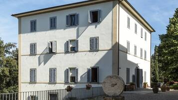 Ein mehrgeschossiges Gebäude mit einer symmetrischen Fassade, zahlreichen Fenstern mit Fensterläden und einem Kieshof mit Topfpflanzen und Außenbestuhlung.