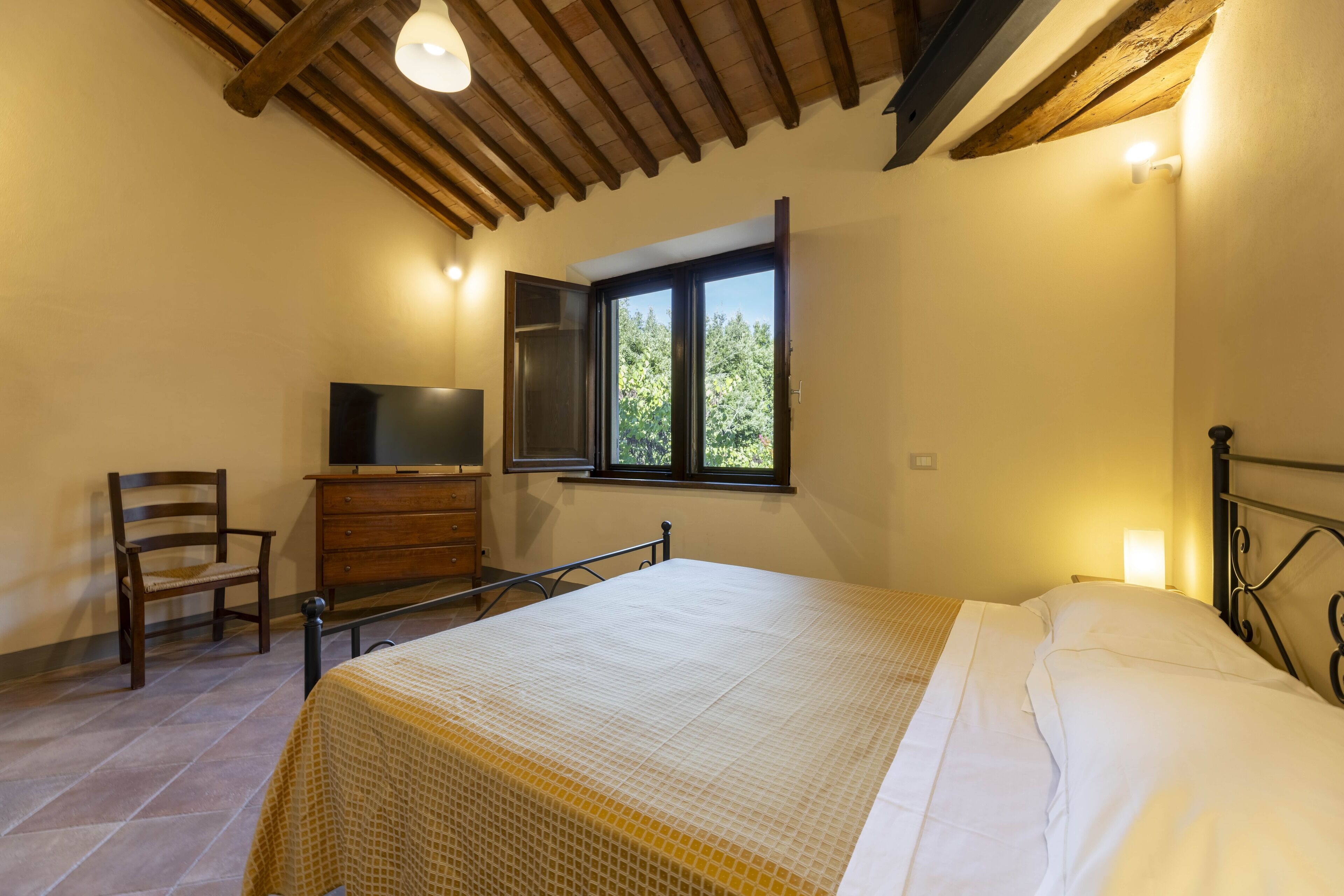 A bedroom with a bed, a dresser, a chair, a television, and a window with a view of trees.