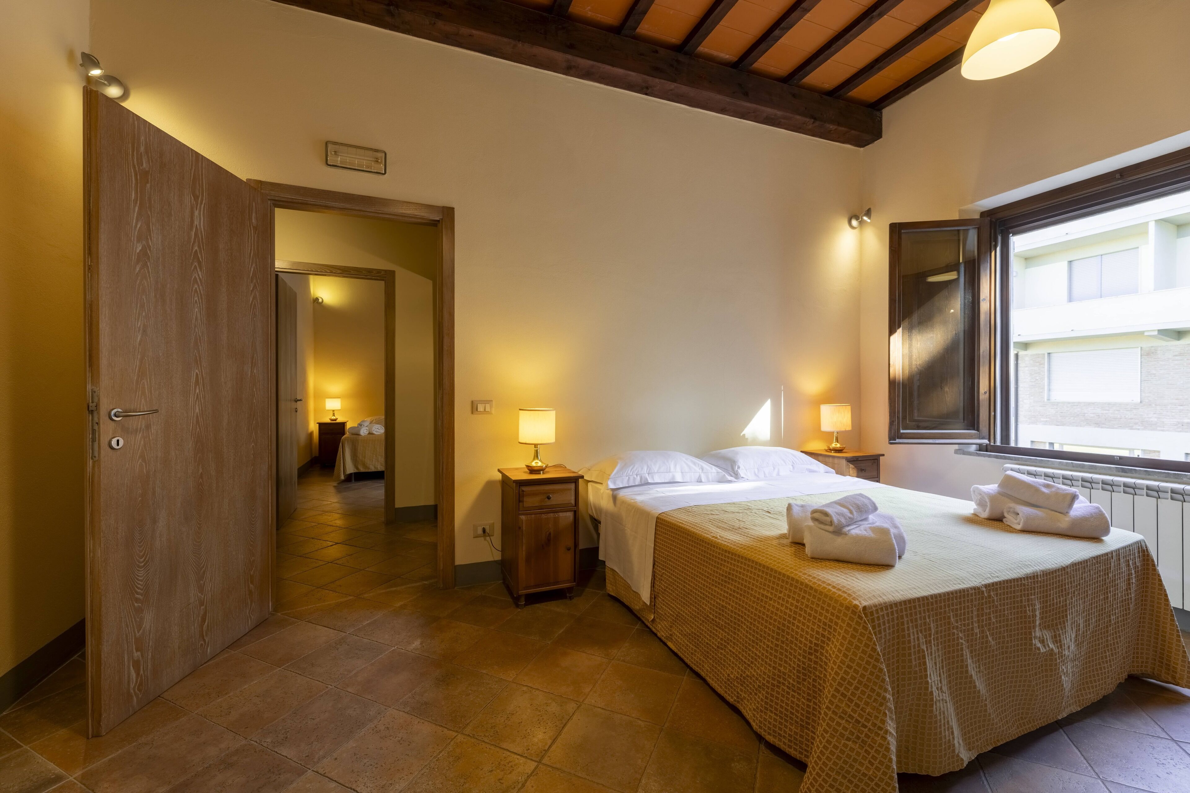 Habitación de hotel con cama, lámparas de noche, ventana y puerta que da a otra habitación.
