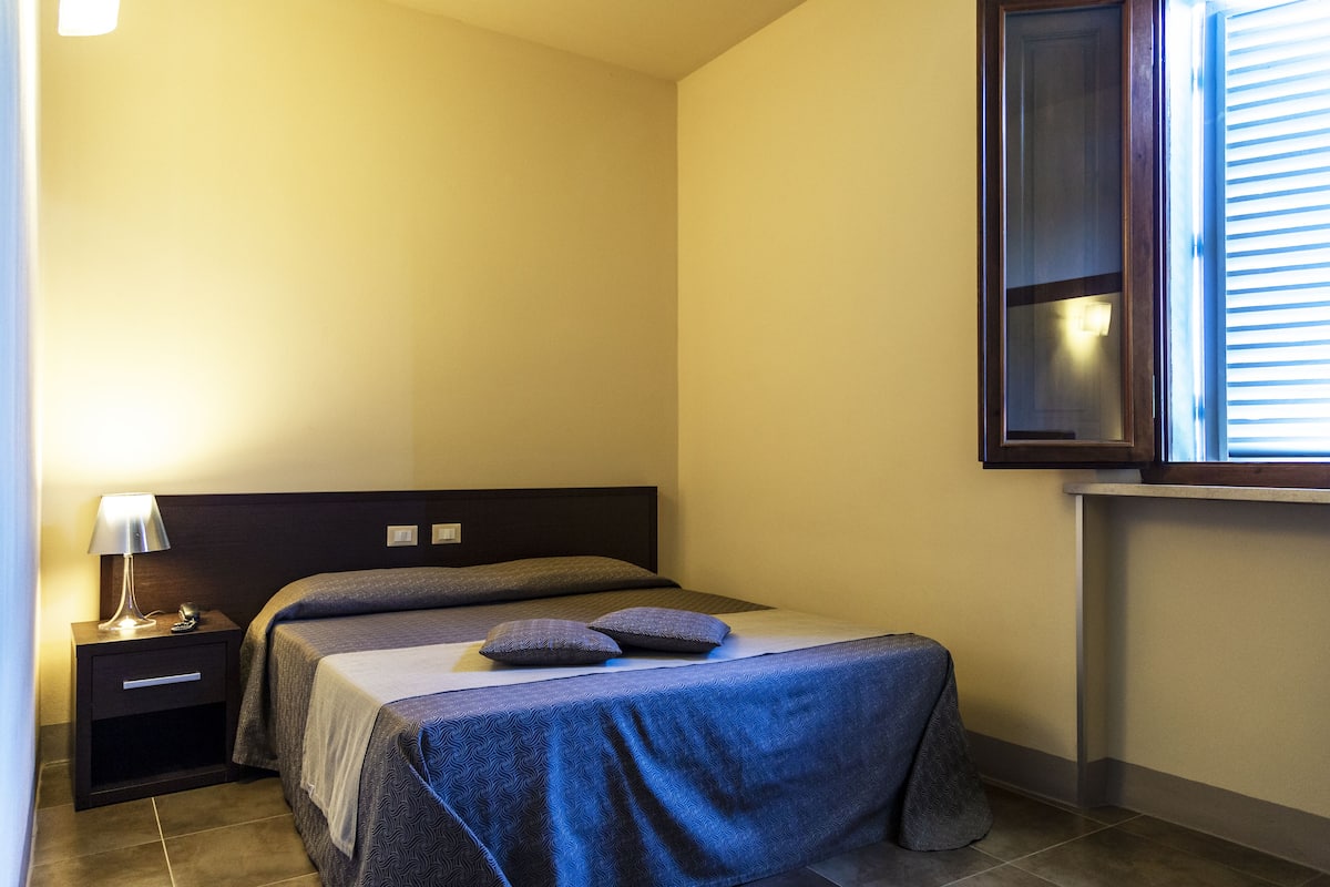 A small, neatly arranged bedroom with a single bed, a nightstand, a lamp, and a window with blinds.