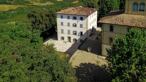 A white building with a tiled roof, surrounded by lush greenery and a hilly landscape. - Villa Montarioso (Monteriggioni)
