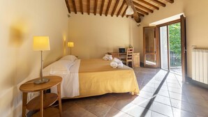 A bedroom with a bed, bedside lamps, a wooden table, and a balcony with a view of greenery. - Villa Montarioso (Monteriggioni)