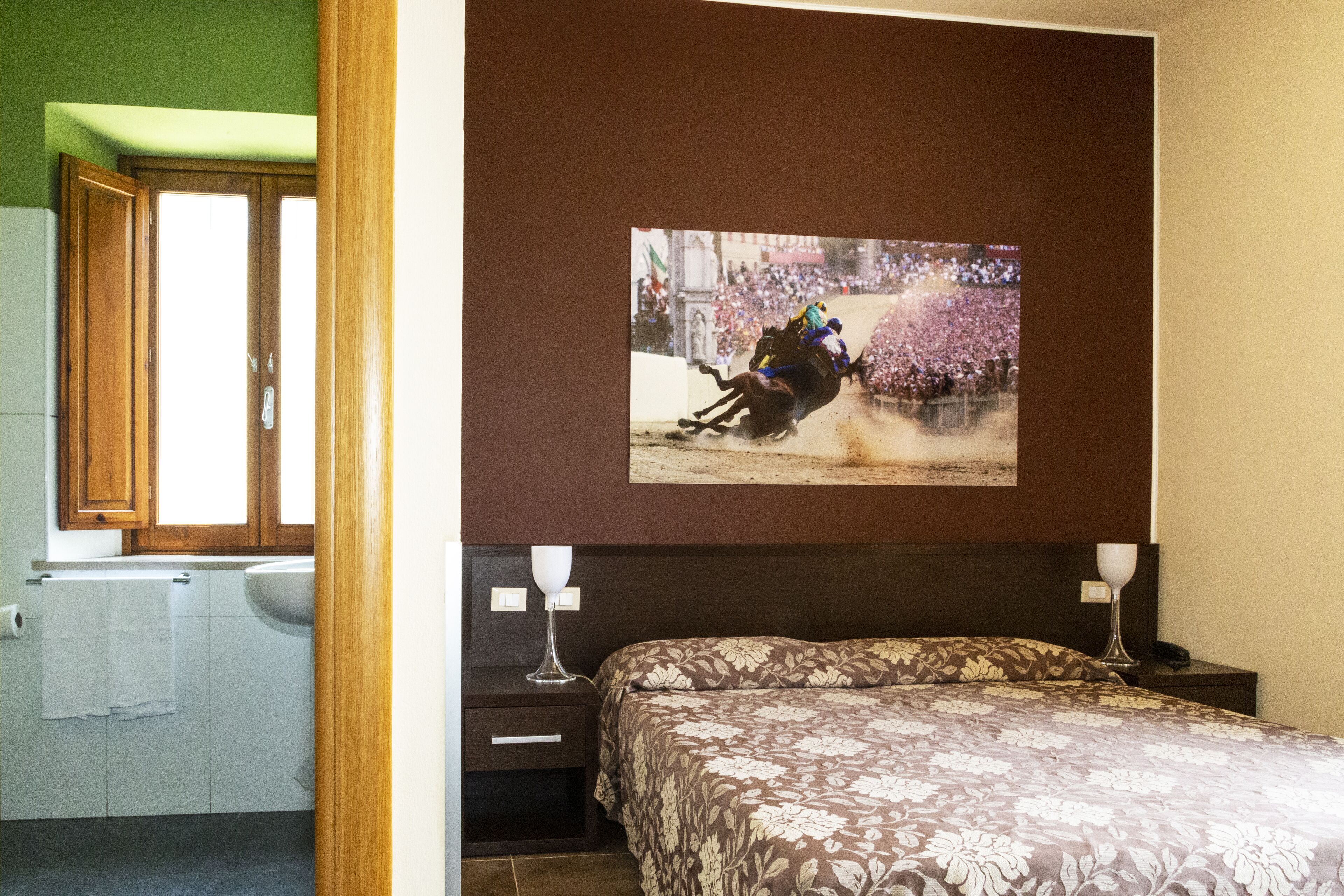 A bedroom with a bed, a nightstand, a wall-mounted TV displaying a horse race, and a bathroom with a toilet and sink.