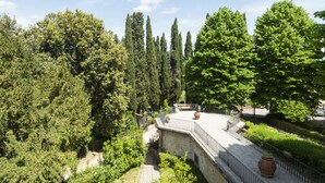 A stone pathway leading to a wooden deck with a railing, surrounded by lush greenery and tall trees. - Villa Montarioso (Monteriggioni)