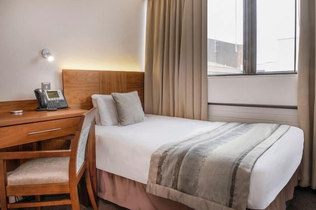 A hotel room with a bed, a desk with a telephone, and a window with curtains.