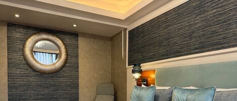 A modern hotel room with a large bed, a round mirror, a chair, and a headboard with multiple pillows.