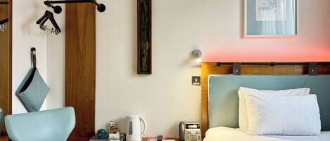 Une chambre d’hôtel avec un lit, une chaise, un bureau avec une bouilloire, un téléphone et un tableau au mur.