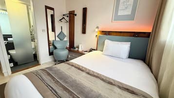 Habitación de hotel con una cama grande, un escritorio con silla, una mesita de noche con lámpara, un perchero y un cuadro en la pared.