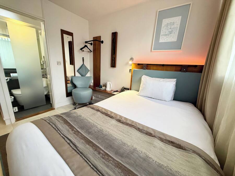 Habitación de hotel con una cama grande, un escritorio con silla, una mesita de noche con lámpara, un perchero y un cuadro en la pared.