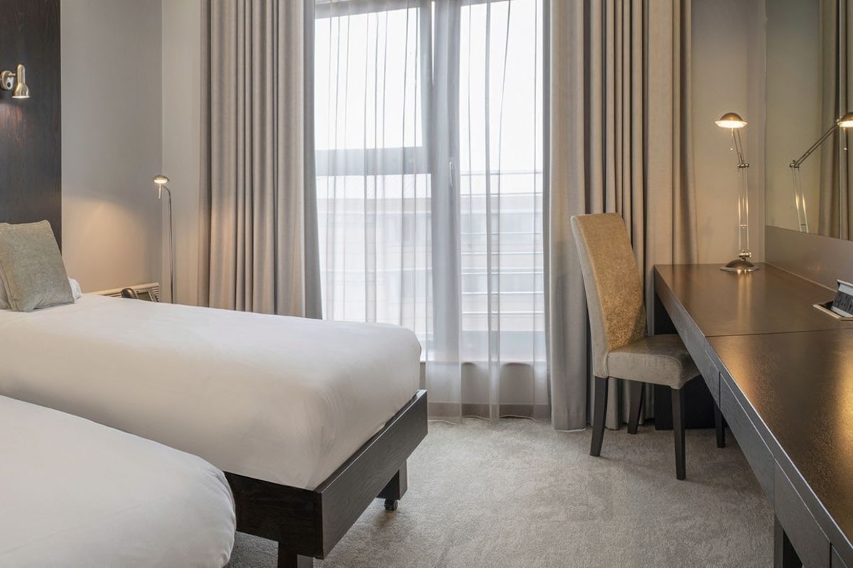 Une chambre d’hôtel avec deux lits, un bureau, une chaise, un miroir et une fenêtre avec des rideaux.
