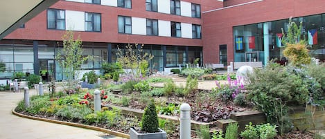 A courtyard with a variety of plants and seating areas, surrounded by a modern building with red brick and large windows.