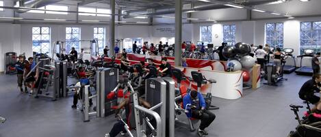 Un gimnasio con diverso equipo de ejercicio, incluyendo cintas de correr y máquinas de pesas, y un ventanal que proporciona luz natural.
