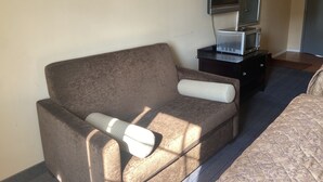 A compact hotel room with a single sofa, a small table with a microwave, a television, and a bed with patterned bedding. - Caravan Inn Motel (Niagara Falls)