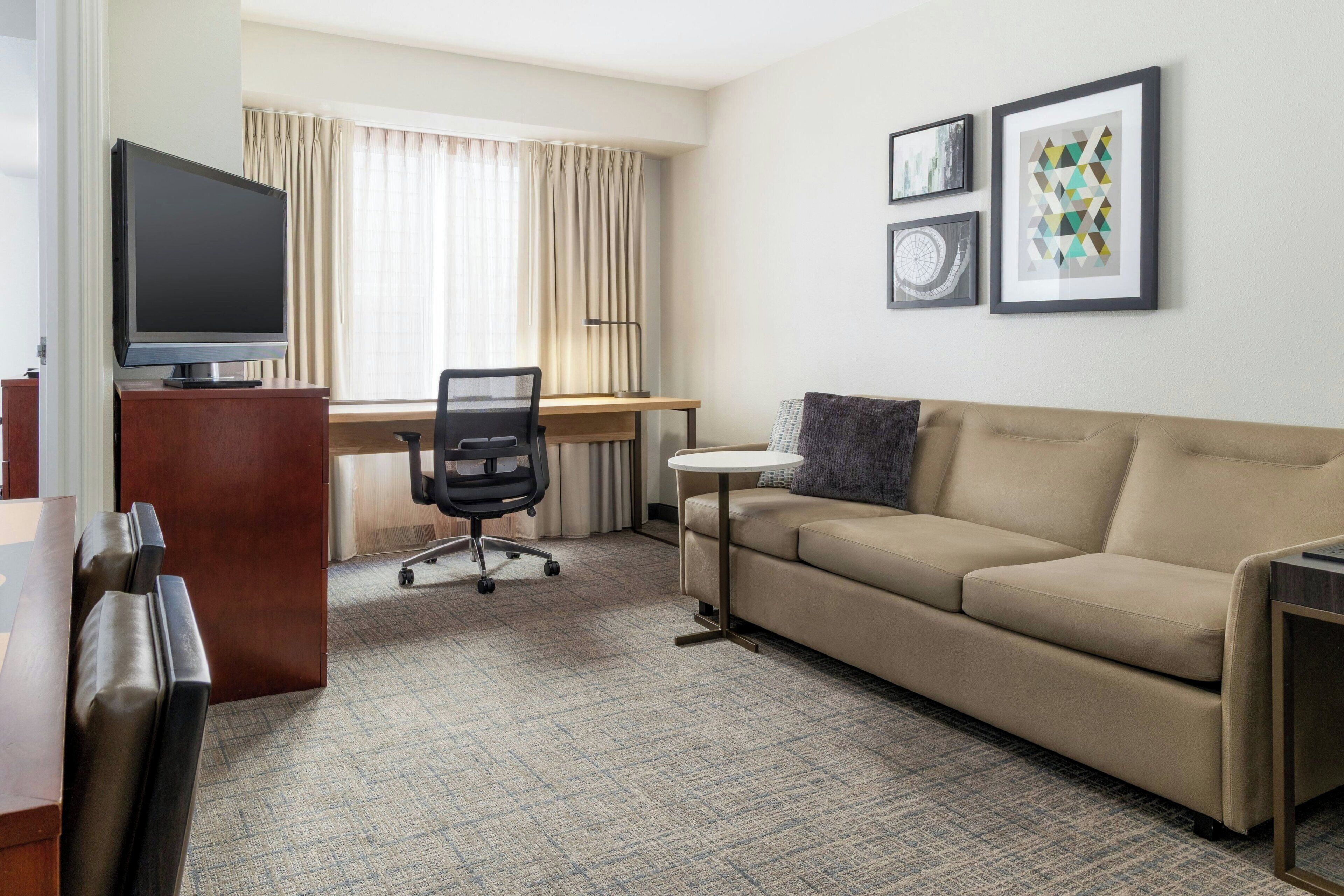 A hotel room with a sofa, a desk, a chair, a television, and framed artwork on the wall.