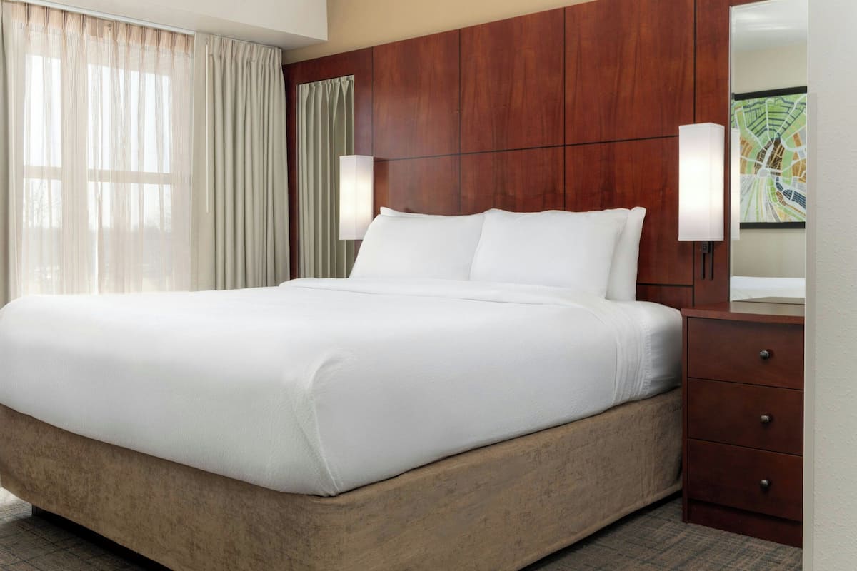 A hotel room with a large bed, wooden headboard, two bedside lamps, a wooden dresser, and a large window with sheer curtains.