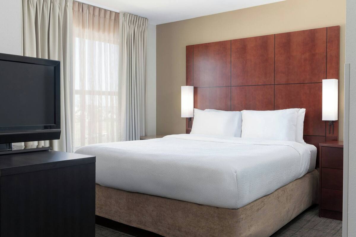 A hotel room with a large bed, a TV on a stand, and two wall-mounted lamps.