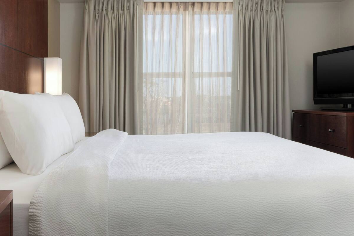 A hotel room with a large bed, a television on a wooden stand, and a window with sheer curtains.