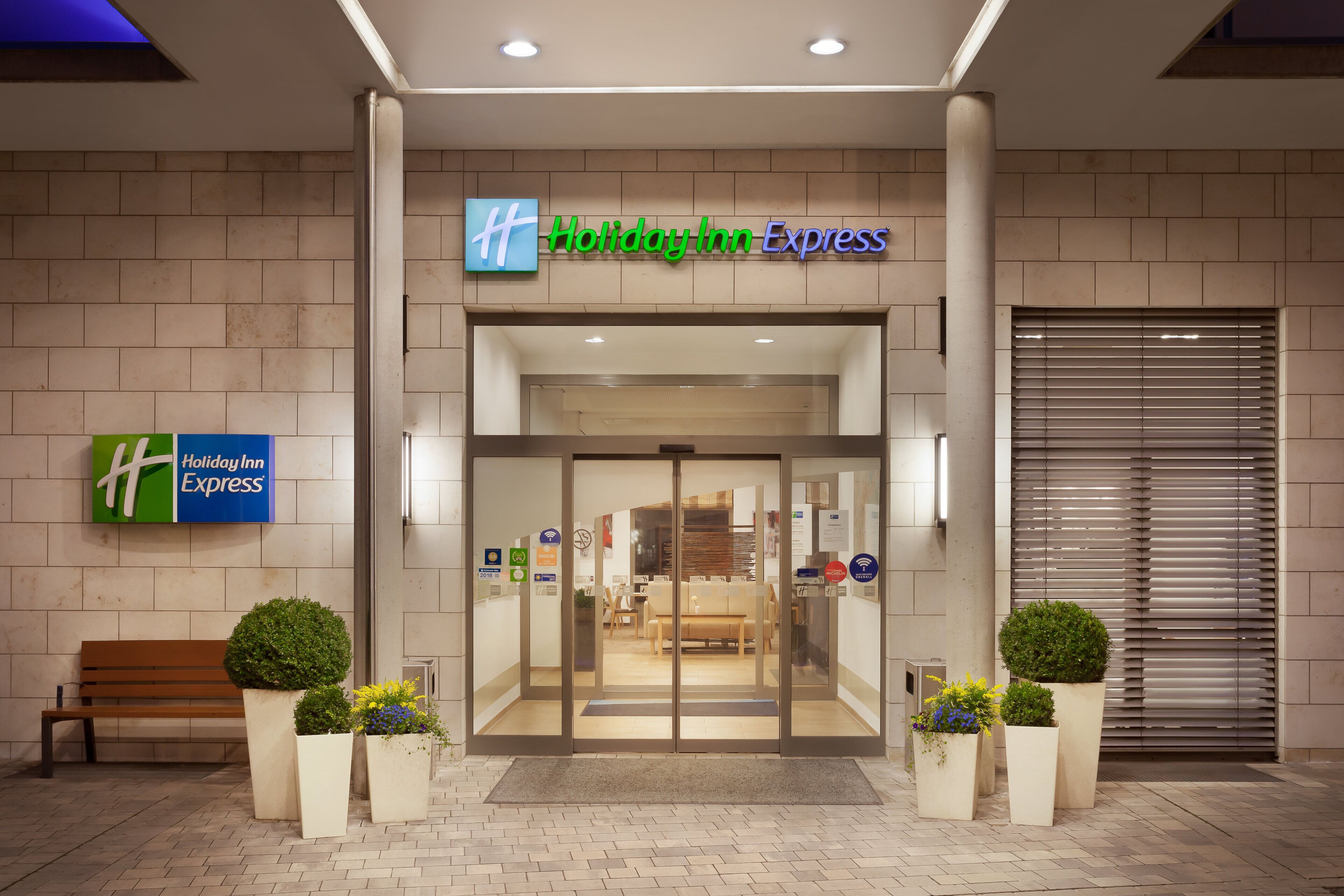 The entrance to a Holiday Inn Express hotel with a modern design, featuring a glass door entrance, a bench, and potted plants.