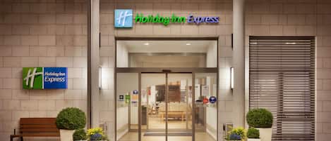 The entrance to a Holiday Inn Express hotel with a modern design, featuring a glass door entrance, a bench, and potted plants.