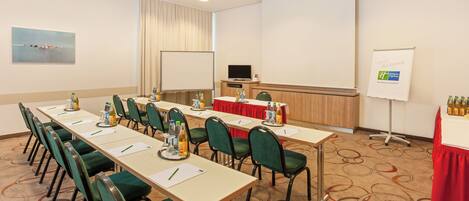 A conference room with a long table, chairs, a projector screen, a whiteboard, and a television.
