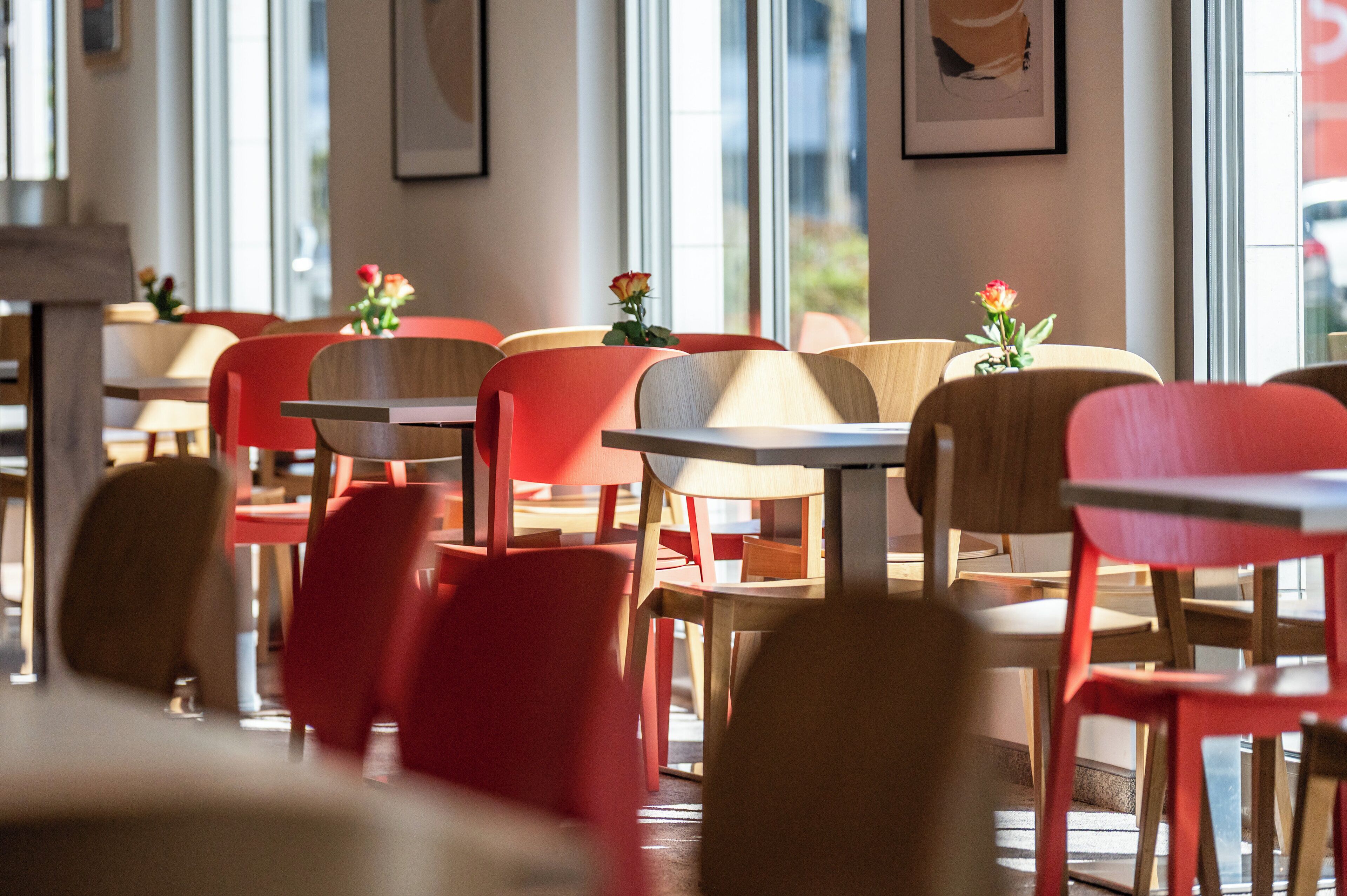 A restaurant interior with red and wooden chairs, tables, and large windows allowing natural light.