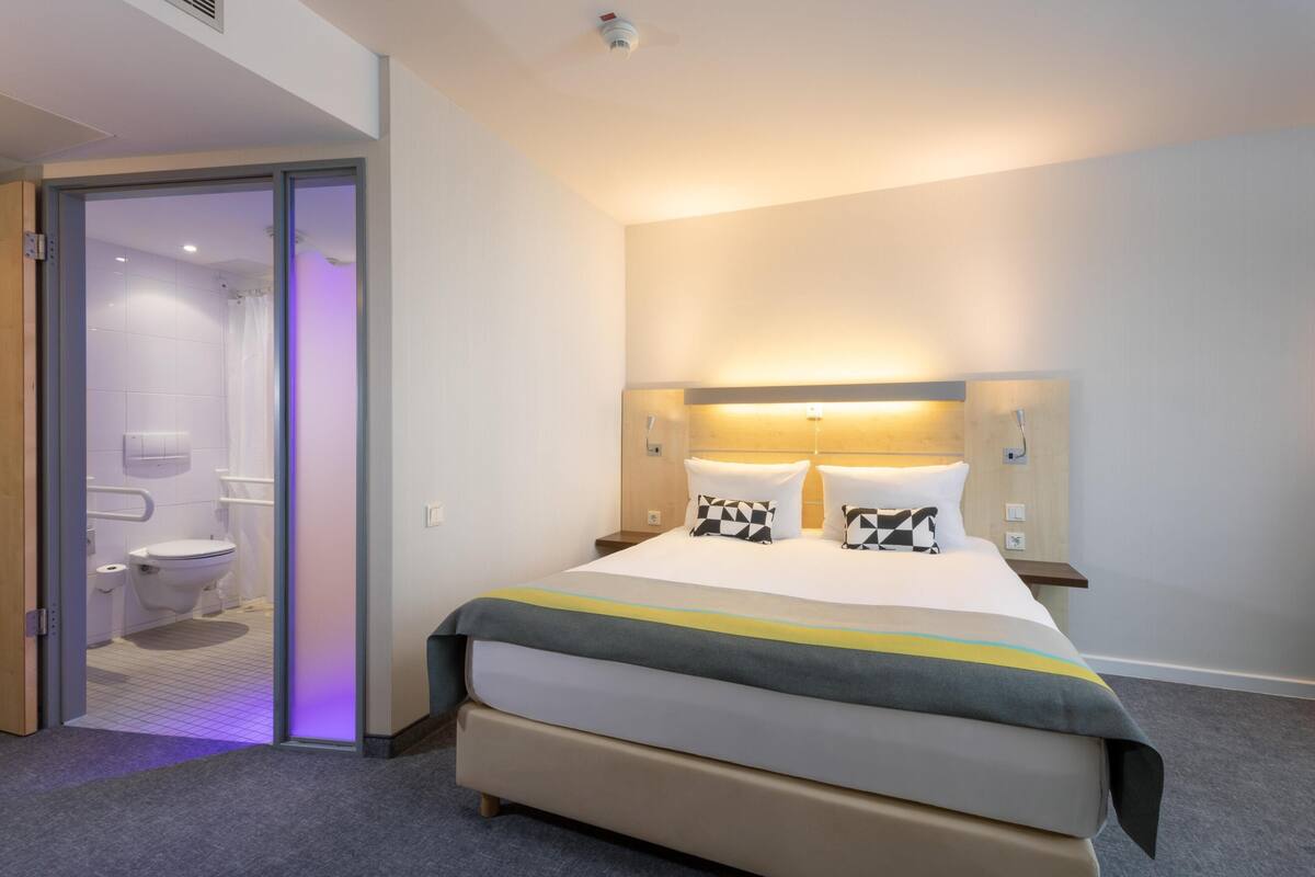 A hotel room with a large bed, a bedside table, a nightstand, a bathroom with a toilet and shower, and a glass door leading to the bathroom.