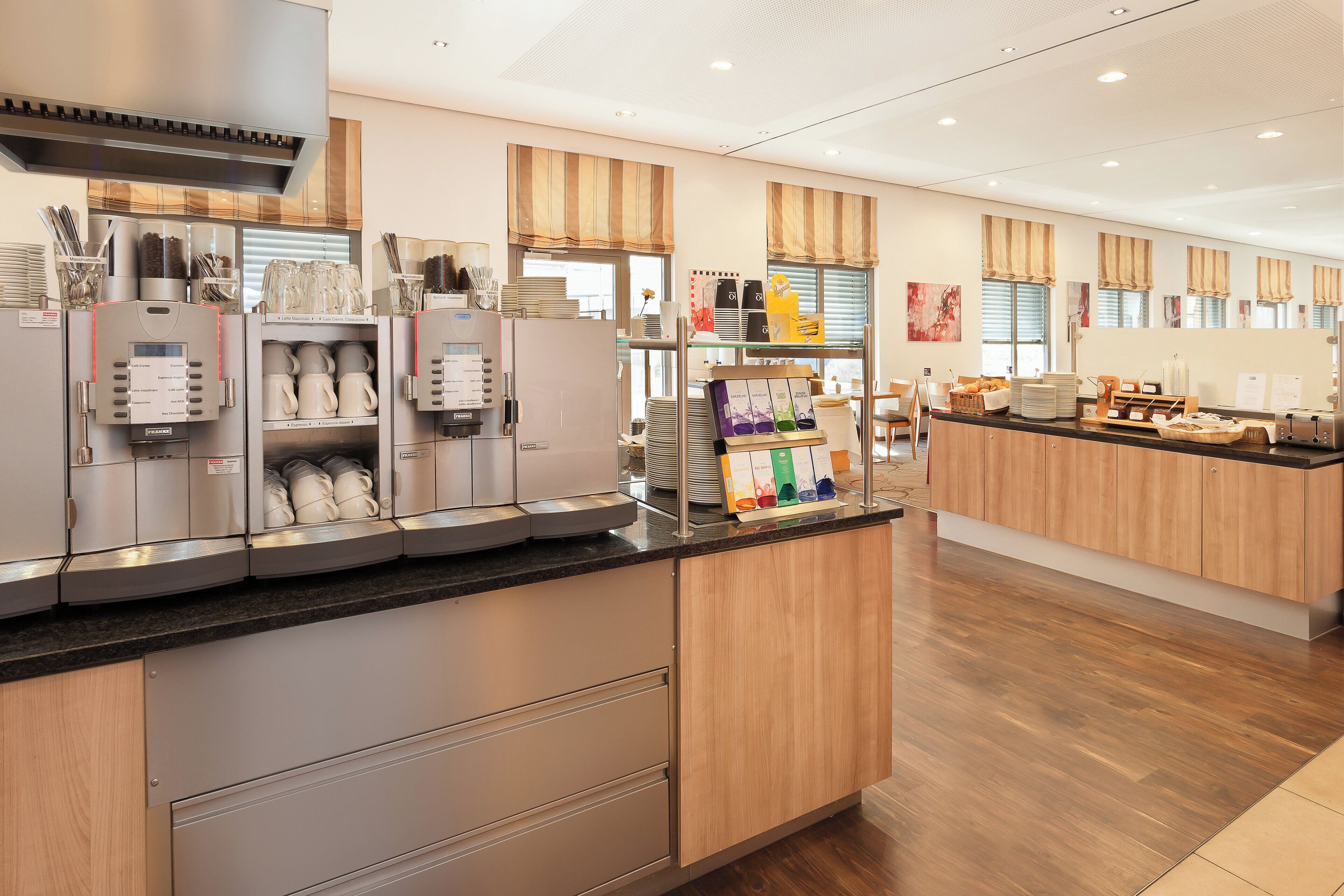 A modern café with a coffee machine, a variety of coffee cups, and a buffet counter with food items.