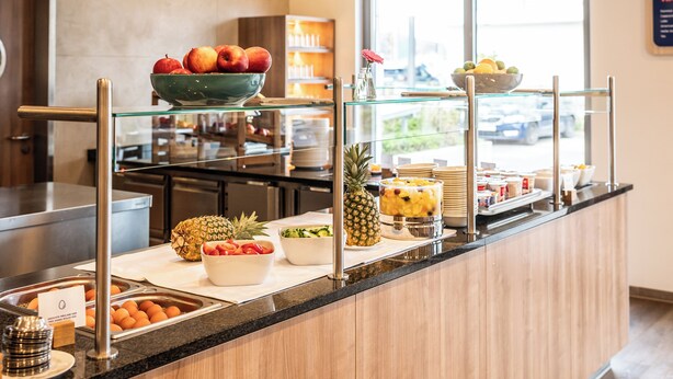 A breakfast buffet with various fruits and dishes, a kitchen area in the background, and a view of the outside through a window. - Holiday Inn Express Nuernberg-Schwabach by IHG (Schwabach)