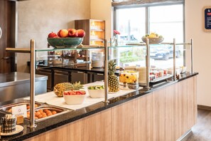 A breakfast buffet with various fruits and dishes, a kitchen area in the background, and a view of the outside through a window. - Holiday Inn Express Nuernberg-Schwabach by IHG (Schwabach)