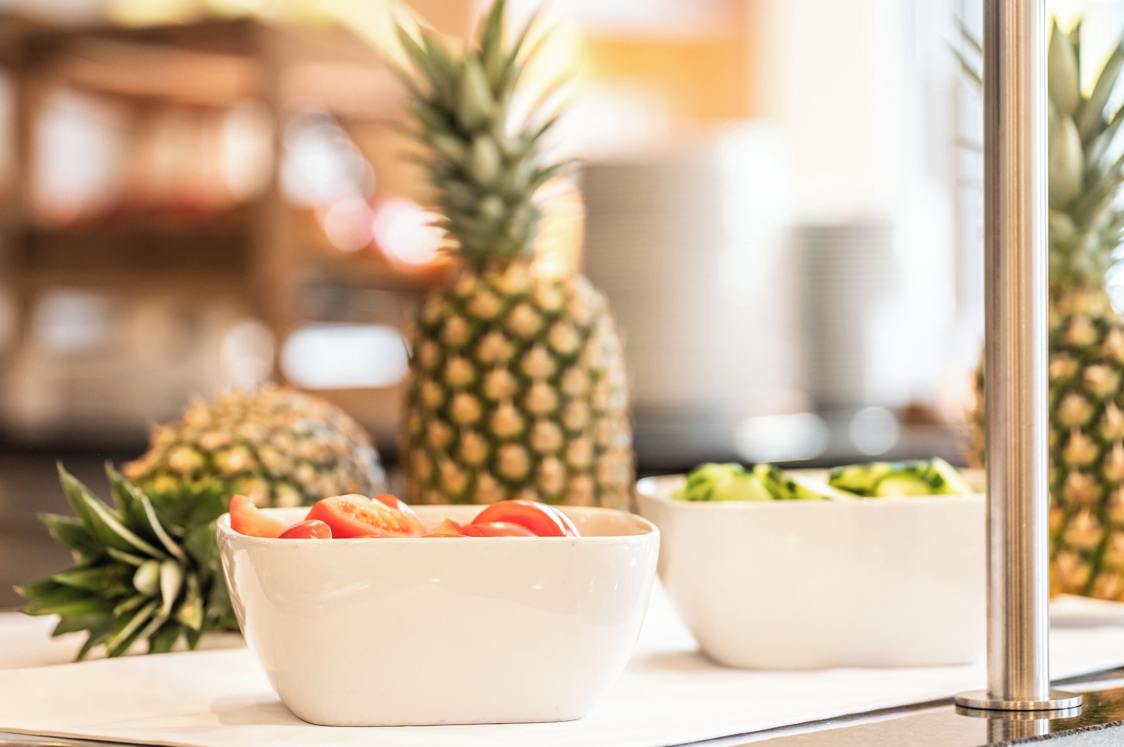 A kitchen counter with a white bowl containing sliced fruit, a white bowl with green vegetables, and two whole pineapples.